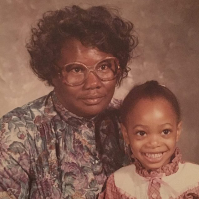 A sepia-colored photo of a Black woman and girl smiling for a portrait against a cloudy brown background. The woman has short, curly black hair and wears brown glasses and a collared shirt with a design of purple flowers. The girl in her lap has braided hair and wears pink shirt with a ruffly white collar and bow.