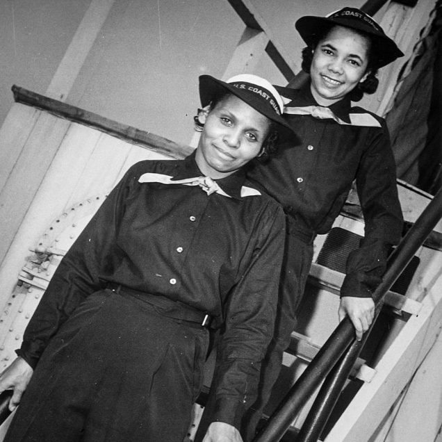 A black-and-white photo of two women, one in front of the other, smiling at the camera on the steps of the ship wearing the U.S. Coast Guard uniform.